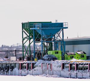 Blue industrial hopper structure with green machinery and vehicles in snowy industrial yard.