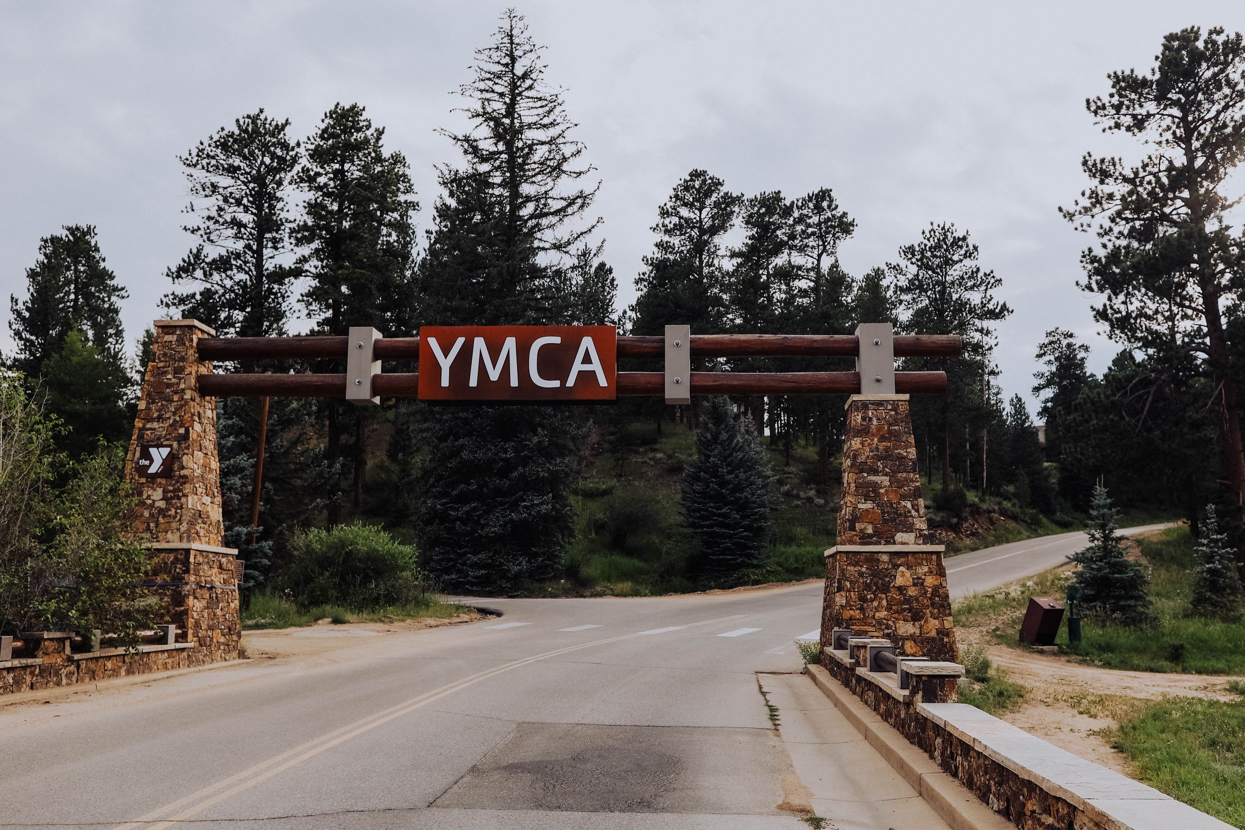 YMLA entrance sign with stone pillars and wooden archway surrounded by pine trees.