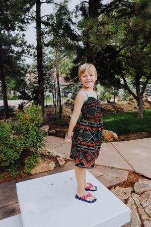 Child jumping off white platform in front of pine trees wearing patterned dress.