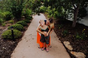Two people in colorful summer outfits embrace while standing on a garden pathway surrounded by landscaping.