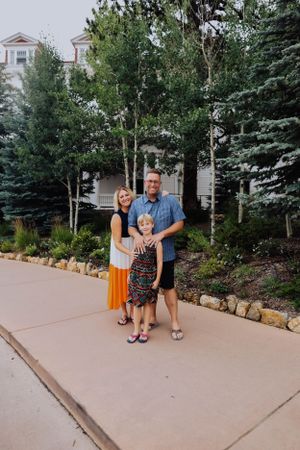 A group poses for a photo on a concrete path with evergreen trees and a building in the background.