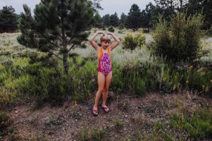 A person in bright swimwear poses with raised arms among desert brush and pine trees.
