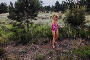 A person in pink swimwear dances playfully in a desert setting with pine trees.