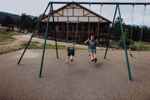Empty playground swings on a grassy field with trees and buildings in the background.