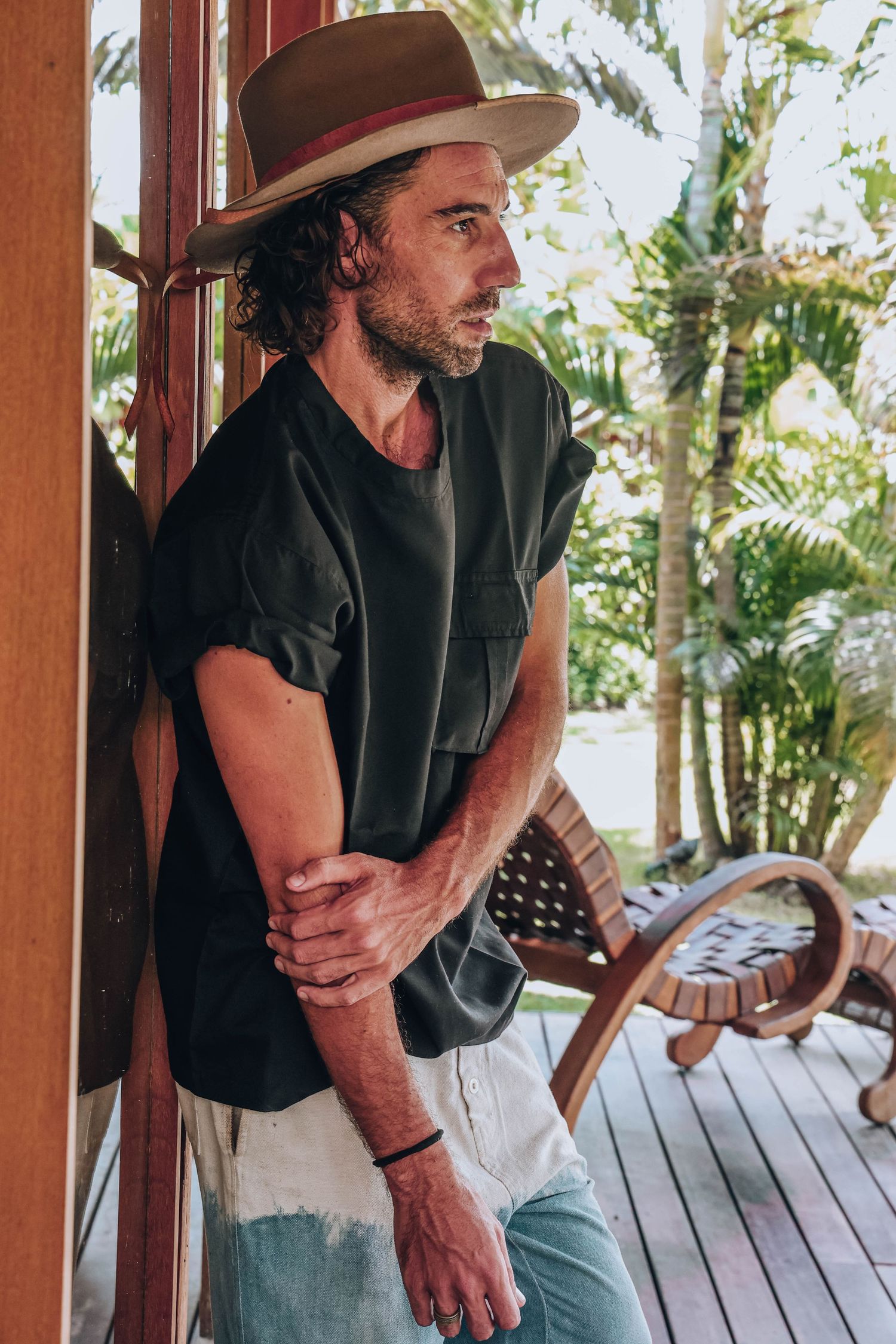 Person in black shirt and hat leaning against wooden post on tropical deck.