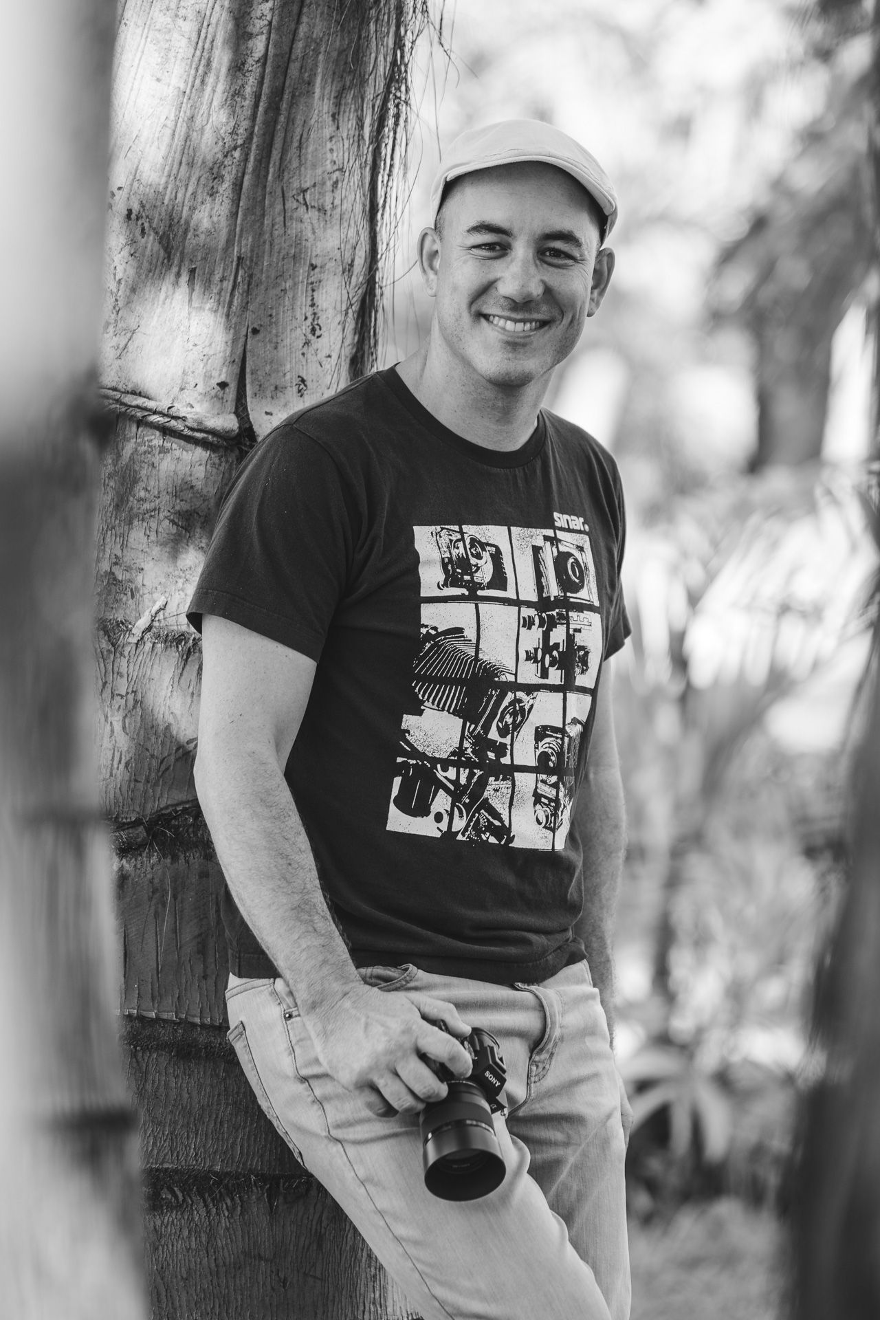 Black and white portrait of photographer in casual attire leaning against wooden post.