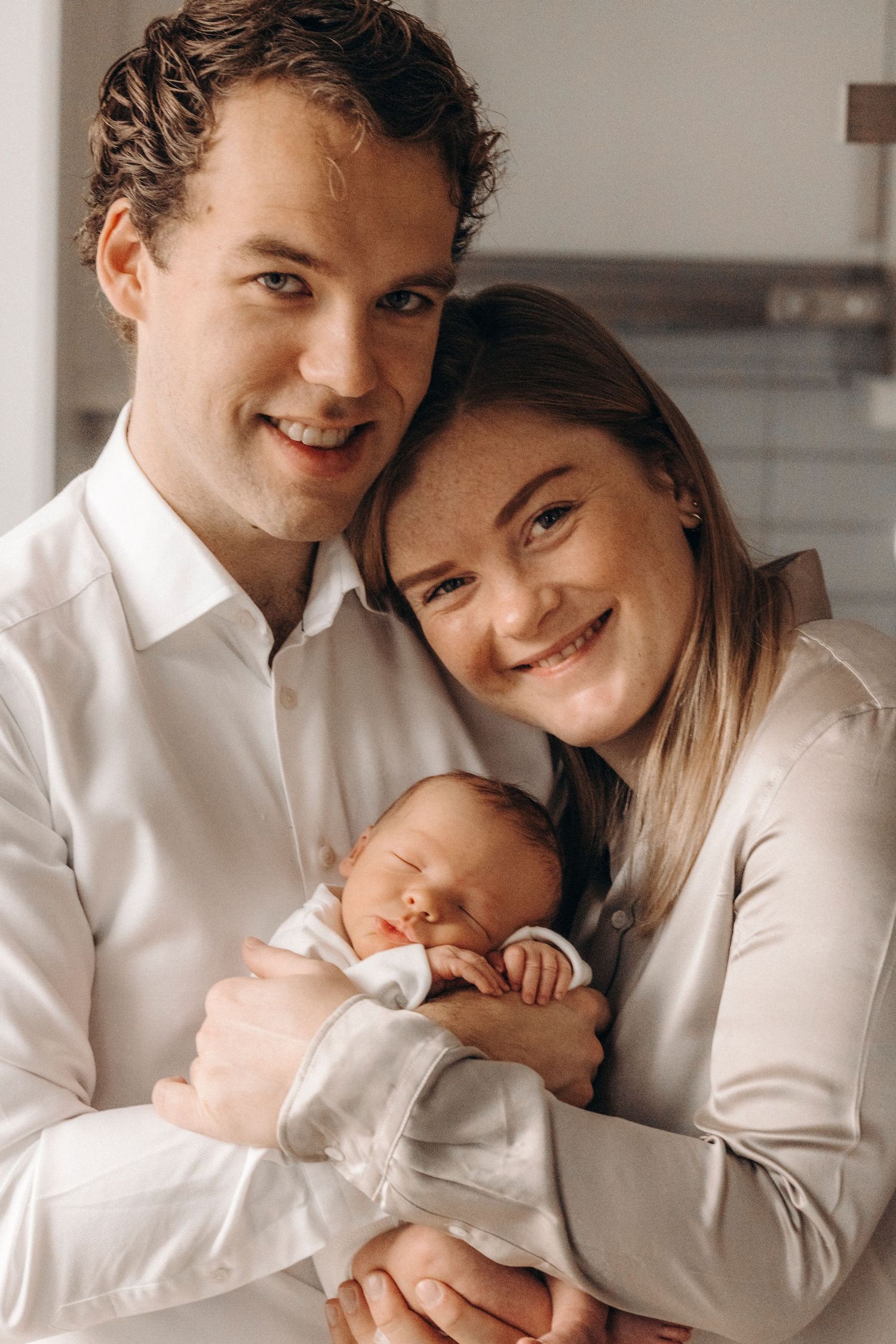 Happy couple in white shirts lovingly holding their sleeping newborn.