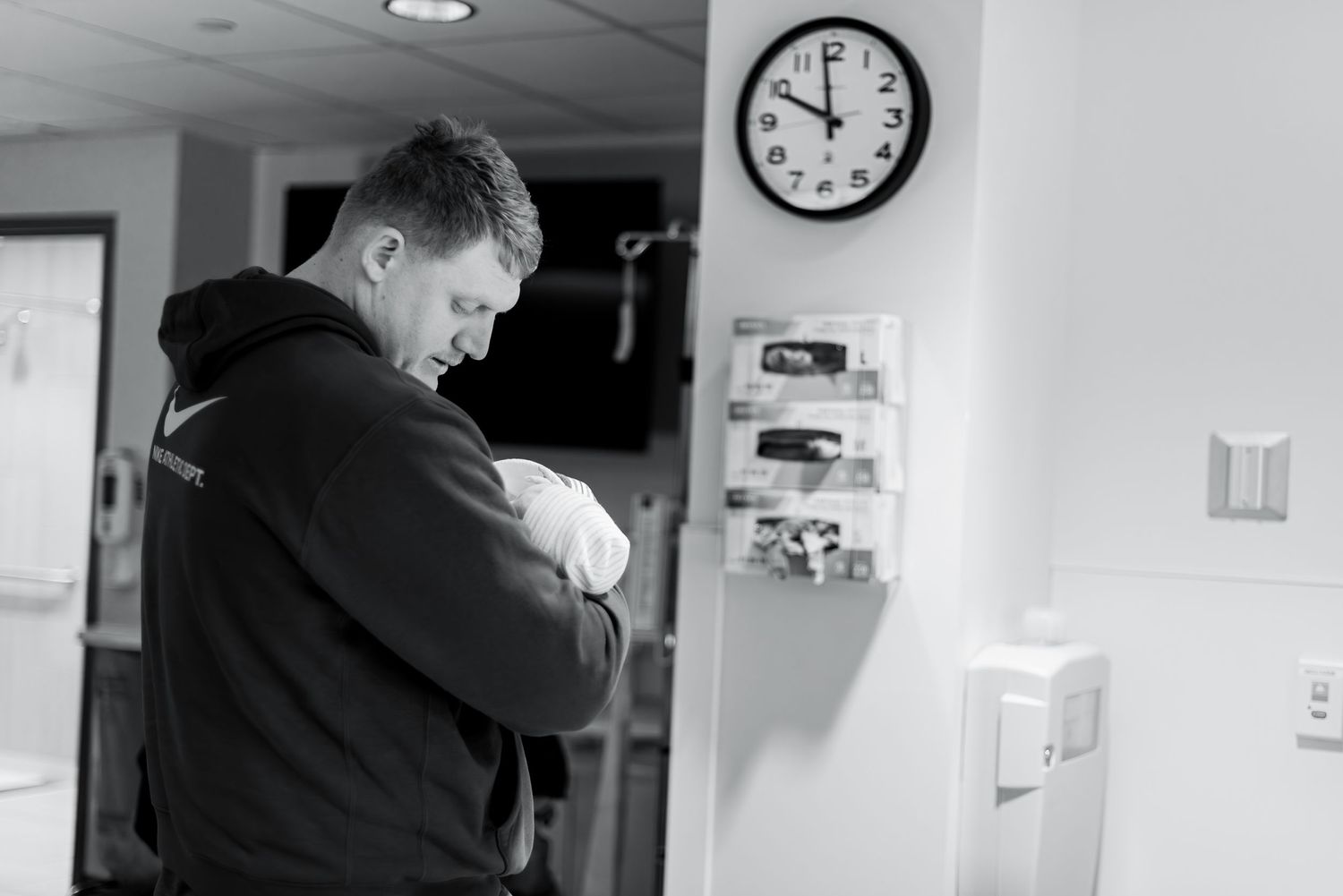 A person in a dark hoodie standing emotionally in a hospital hallway near a wall clock.
