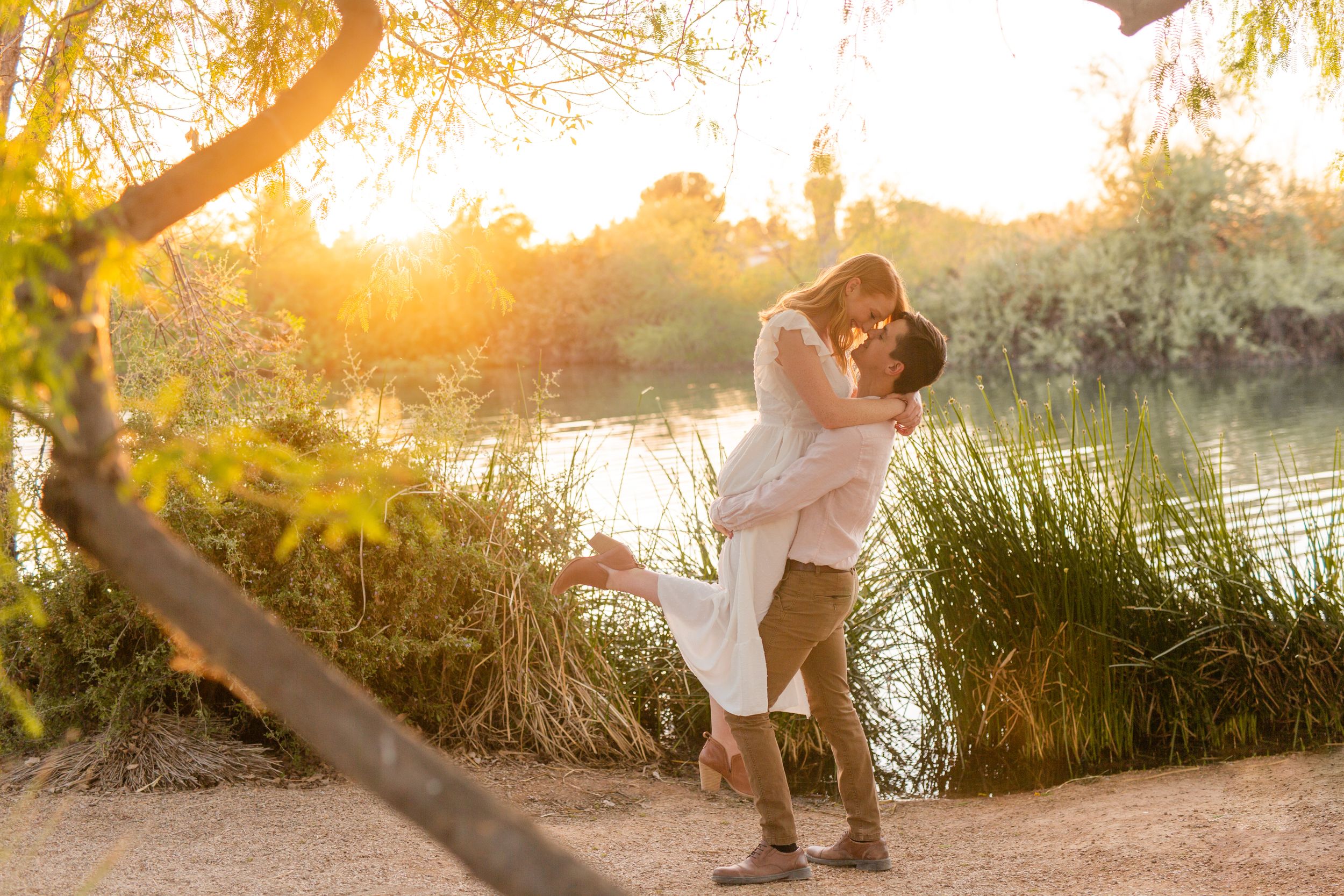 Couple sharing romantic moment at sunset by river with golden light filtering through trees.