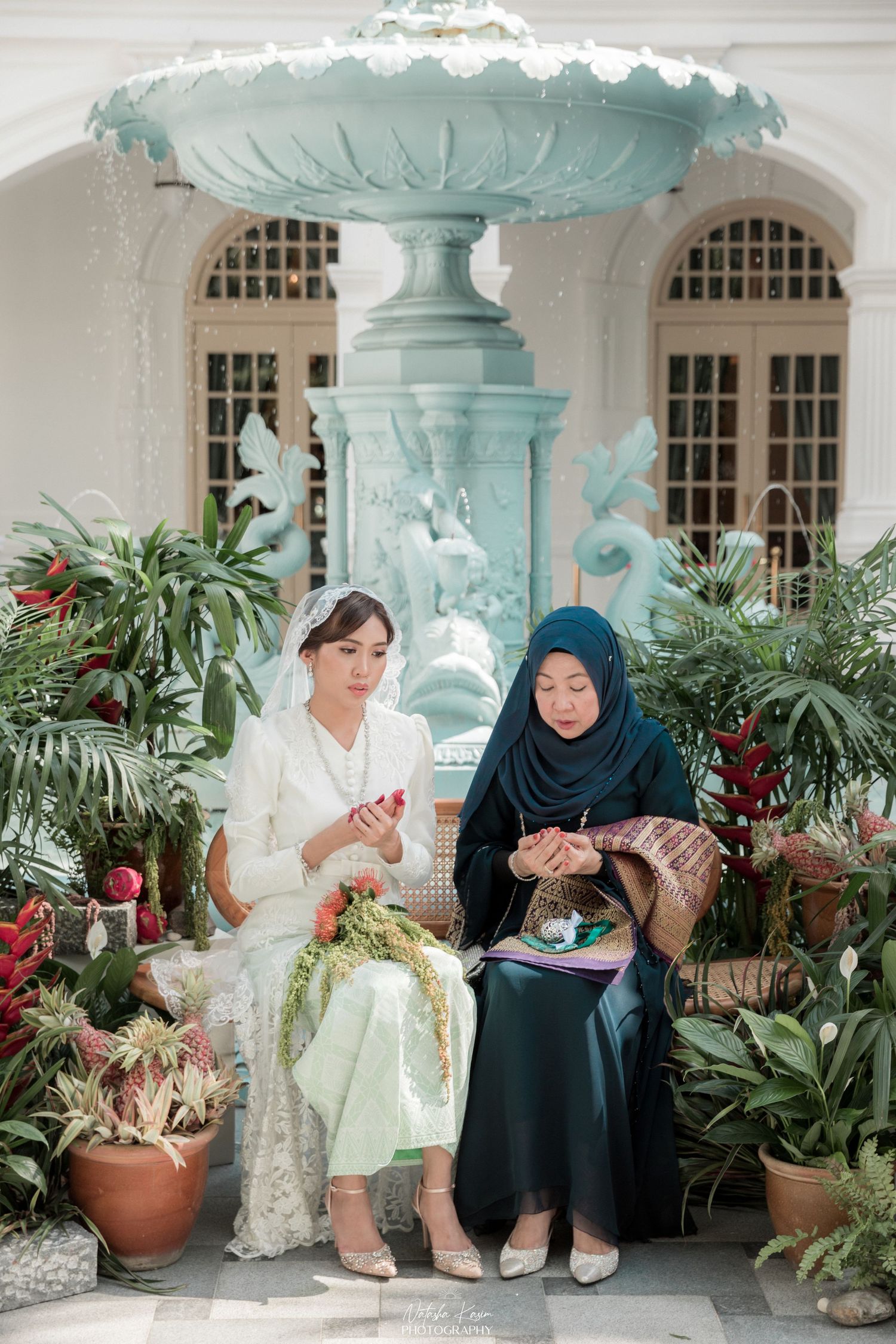 Two women in traditional attire sit on a bench near a decorative mint fountain surrounded by potted plants.