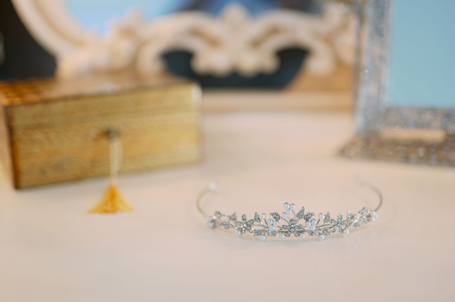 A delicate crystal tiara with floral details rests on a white surface next to a decorative gold box.