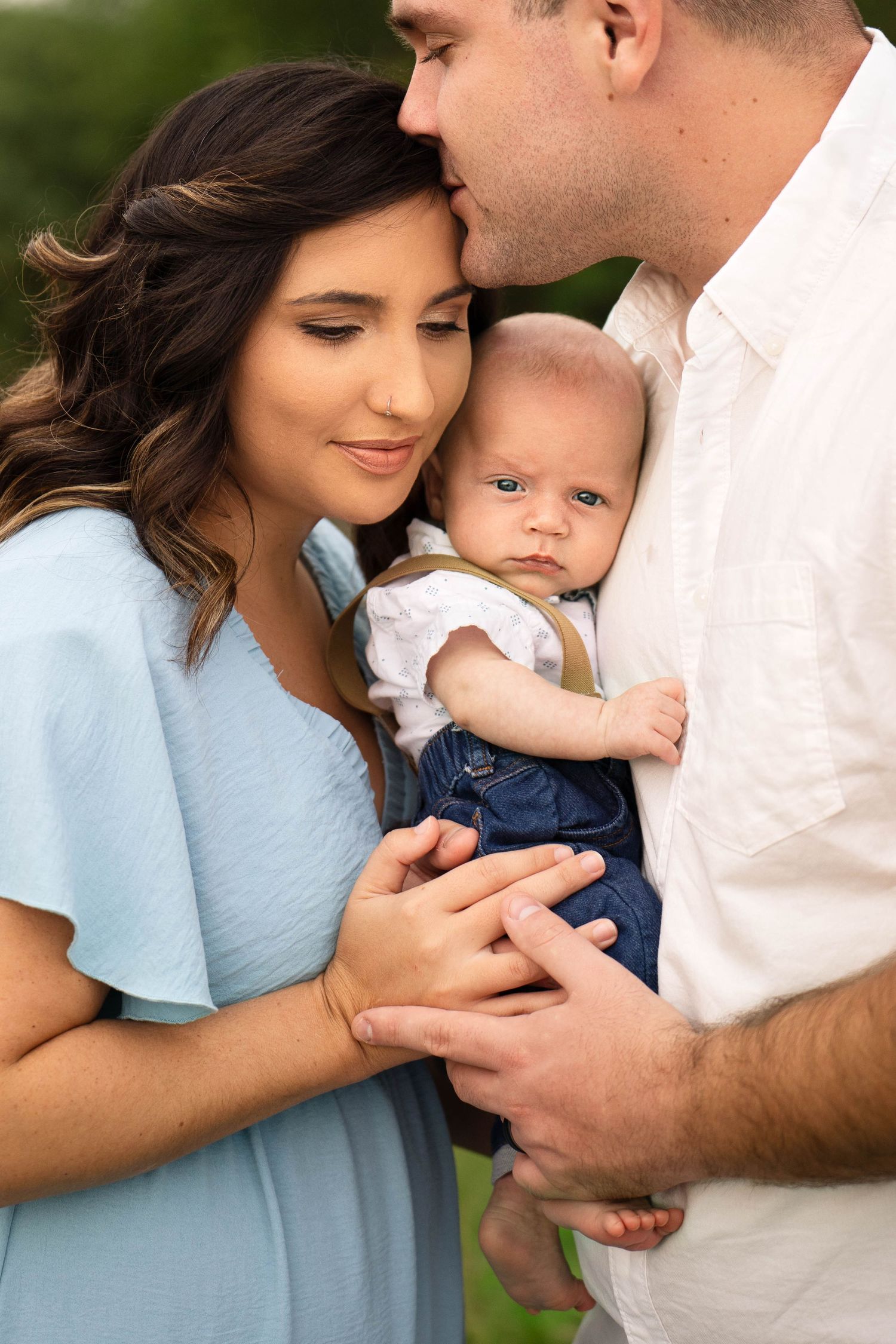 a young husband holds his newborn against his chest as he leans into his wife's forehead and gives her a kiss