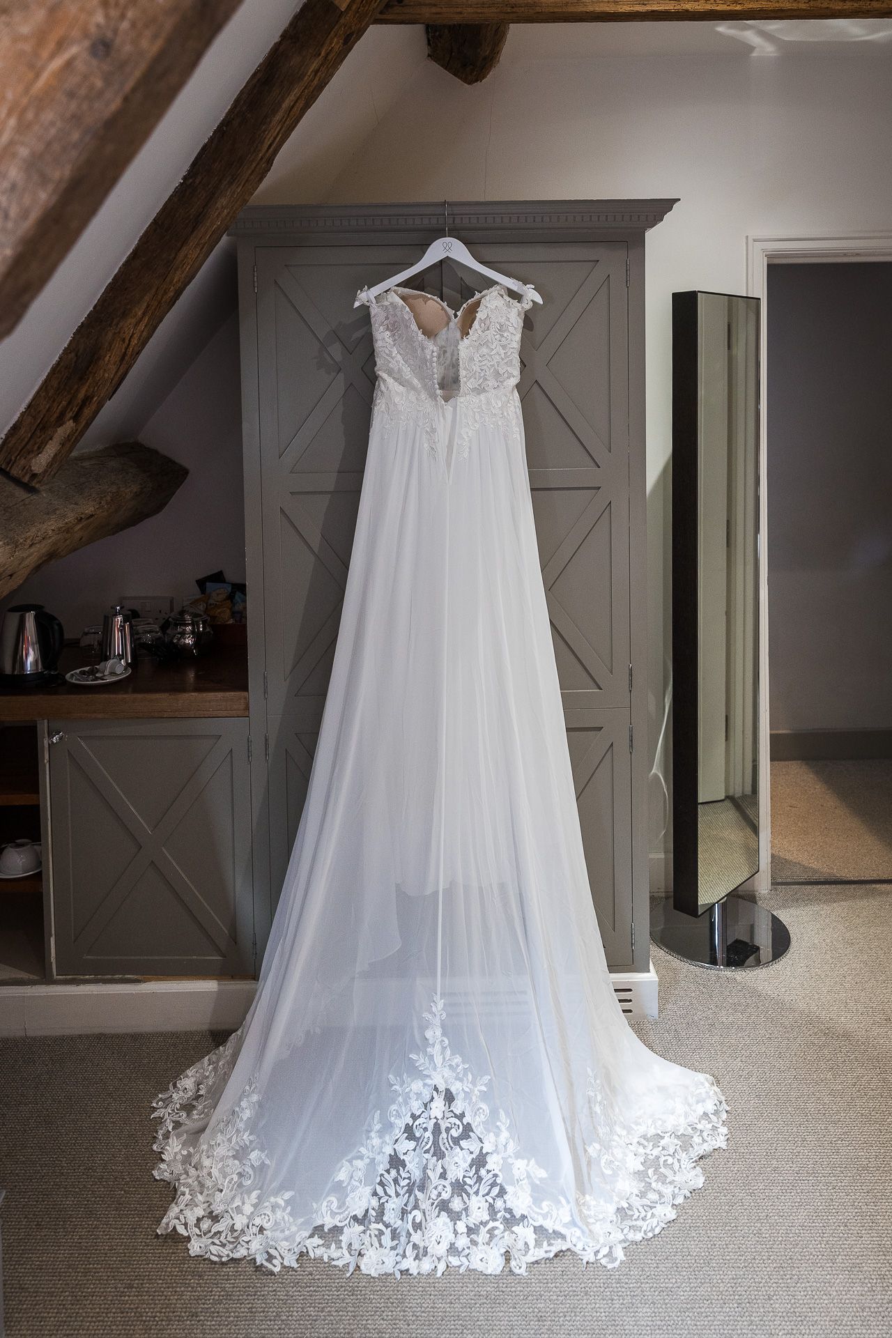 Elegant white lace wedding gown hanging against gray paneled wall in attic room.