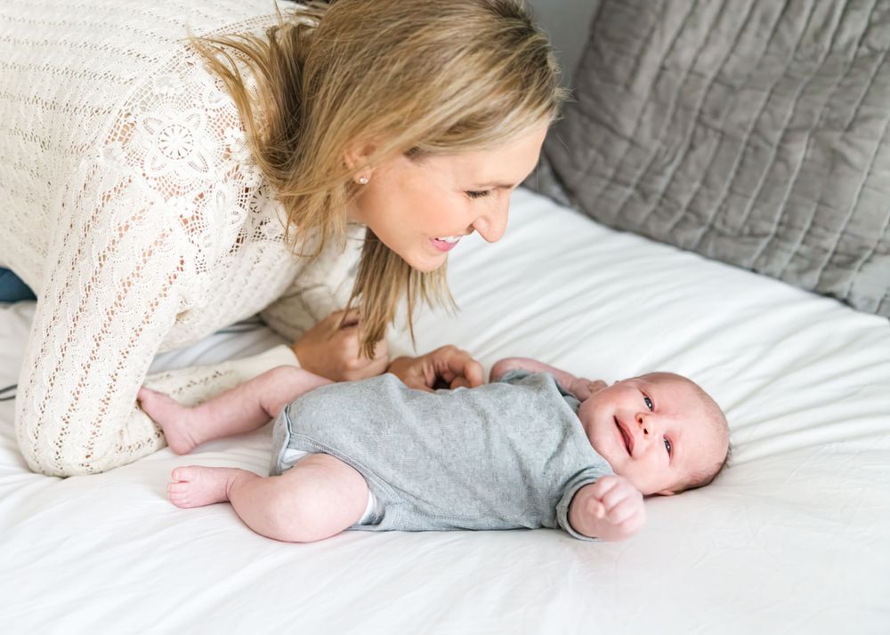Sweet moment between mother and laughing baby in gray onesie lying on white bedding.