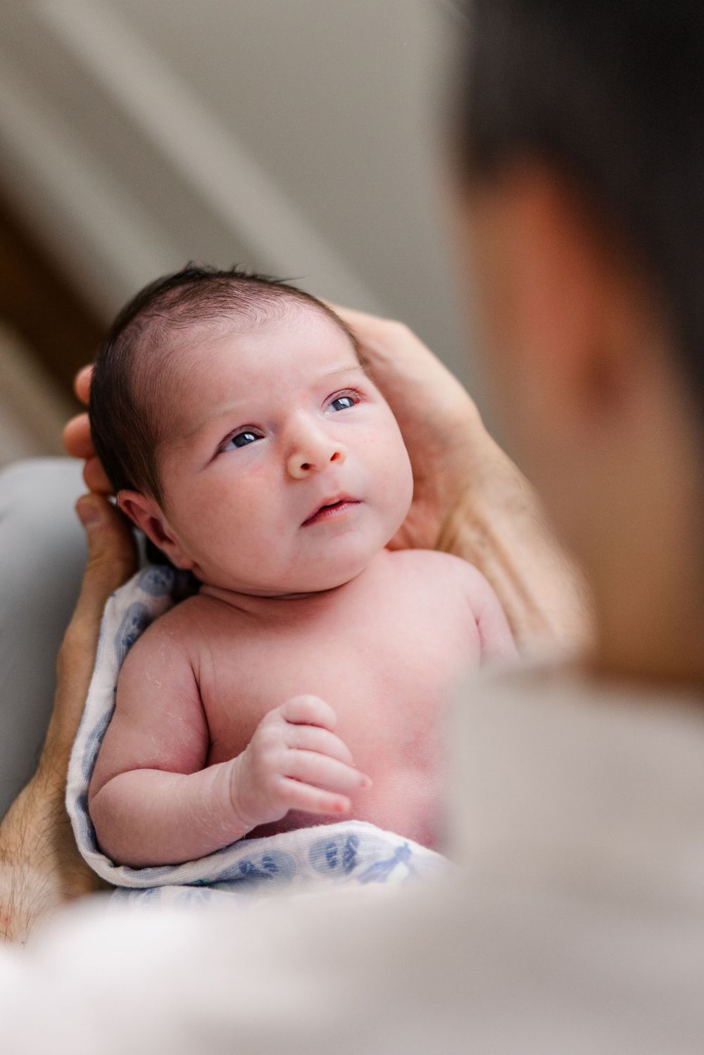 Small baby lies on a blanket while being gently held, looking up with bright eyes in soft lighting.