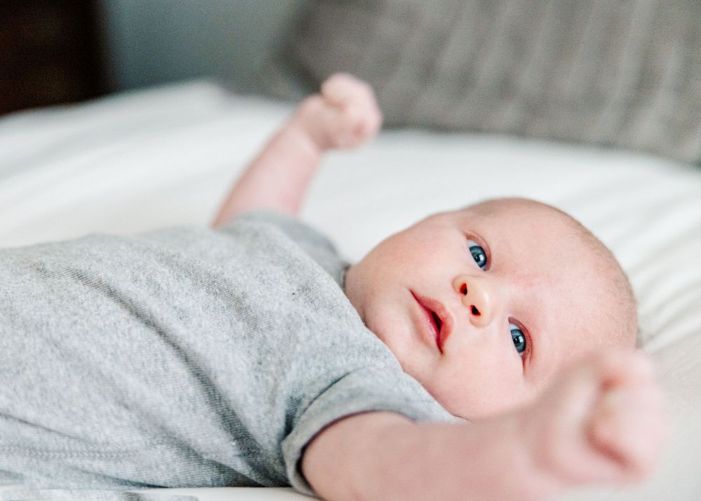 Baby wearing gray sleeper lies on white bedding while gazing upward in a thoughtful moment.