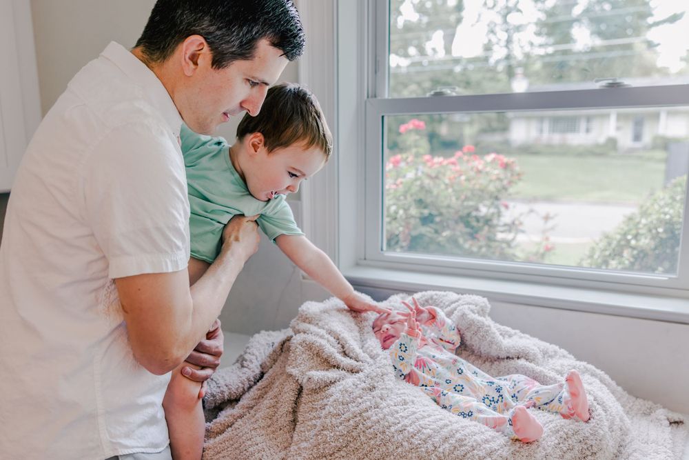 A father and small child look down at a newborn baby wrapped in floral blankets by a sunny window.