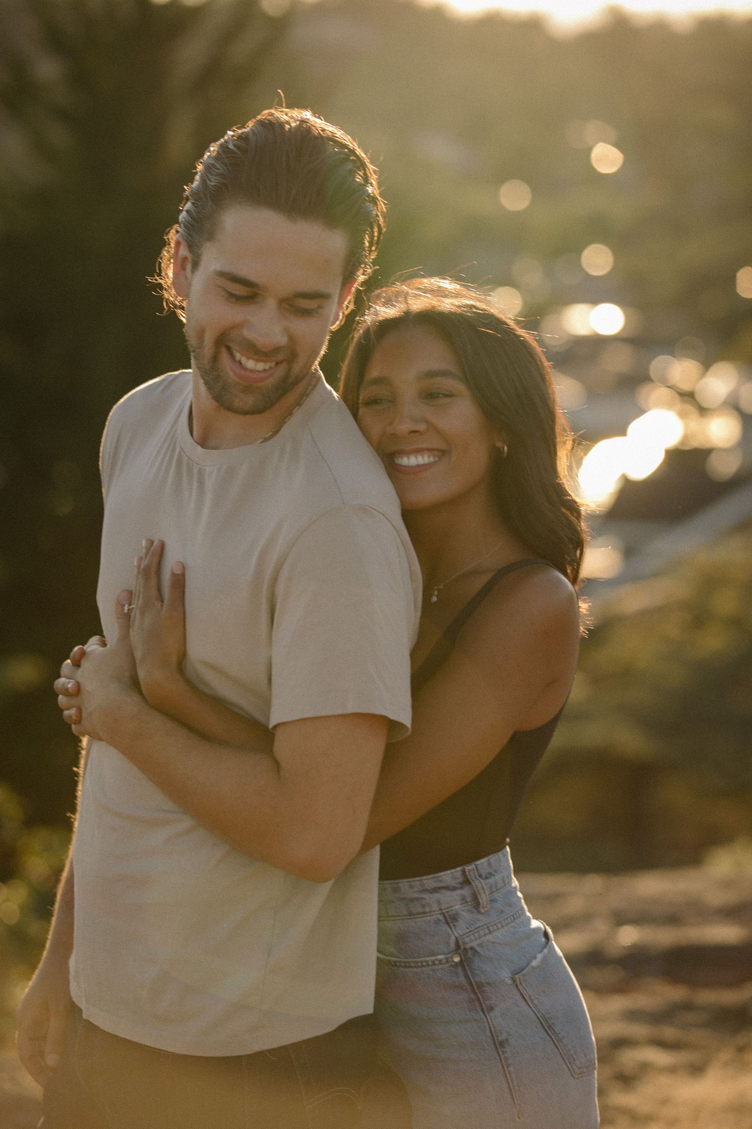 Girlfriend holds boyfriend at sunset while he smiles back at her
