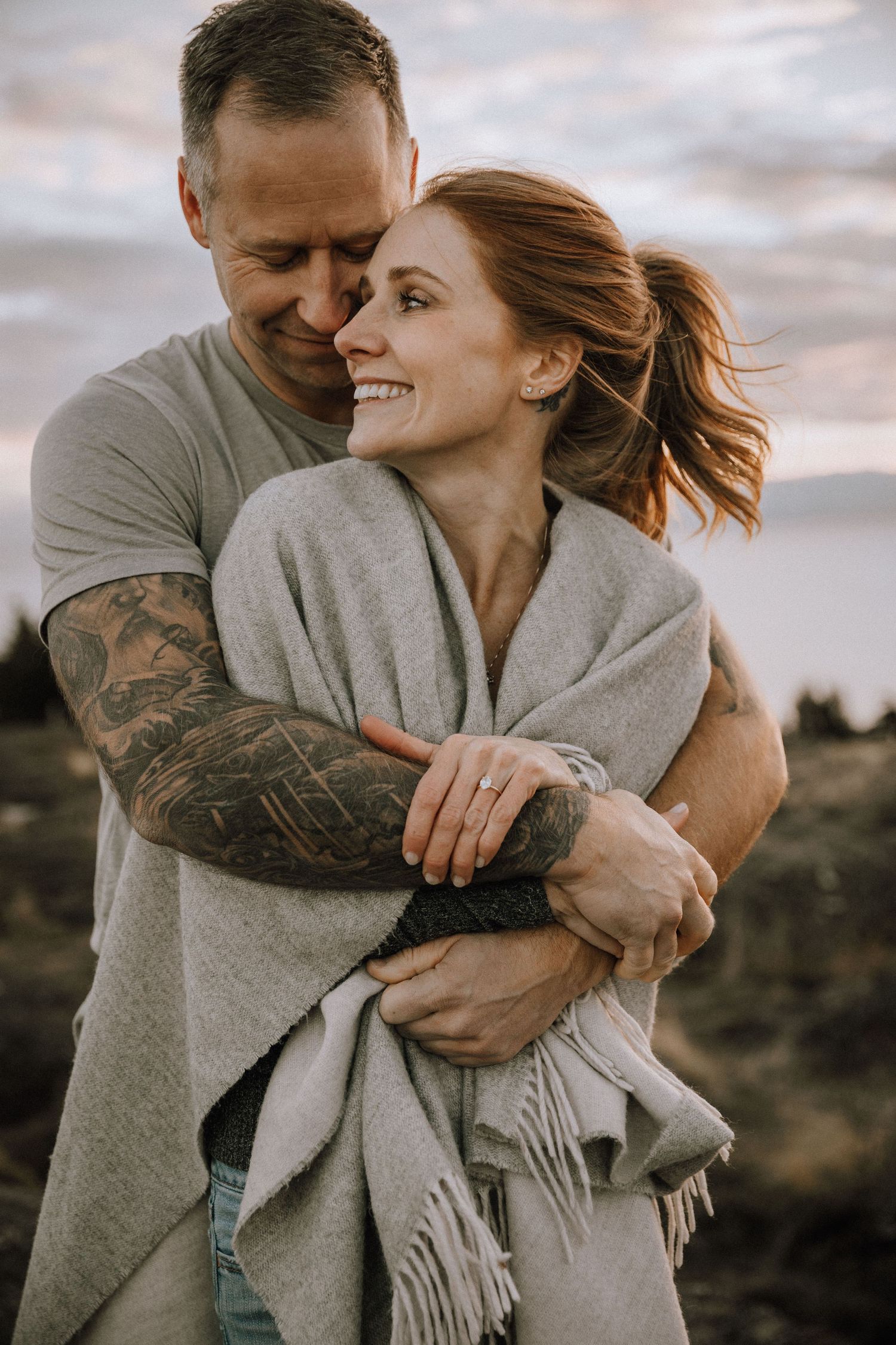 Boyfriend bear hugs girlfriend with a blanket wrapped around her at sunset