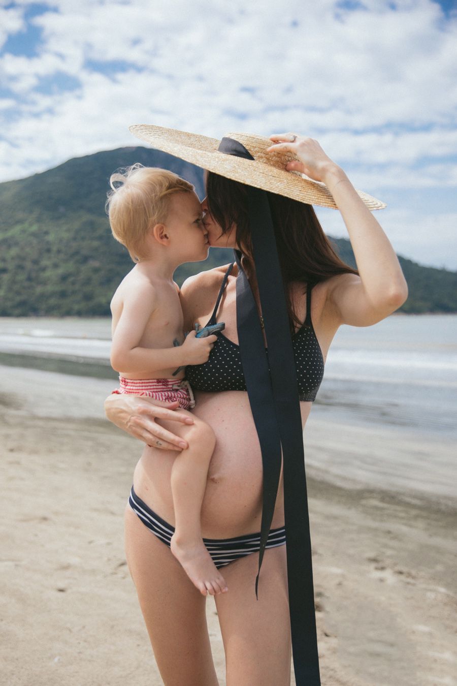 Pregnant mom holds young son on the beach as they kiss