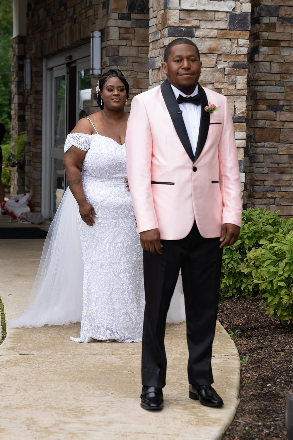 A couple in wedding attire pose outside a stone building.