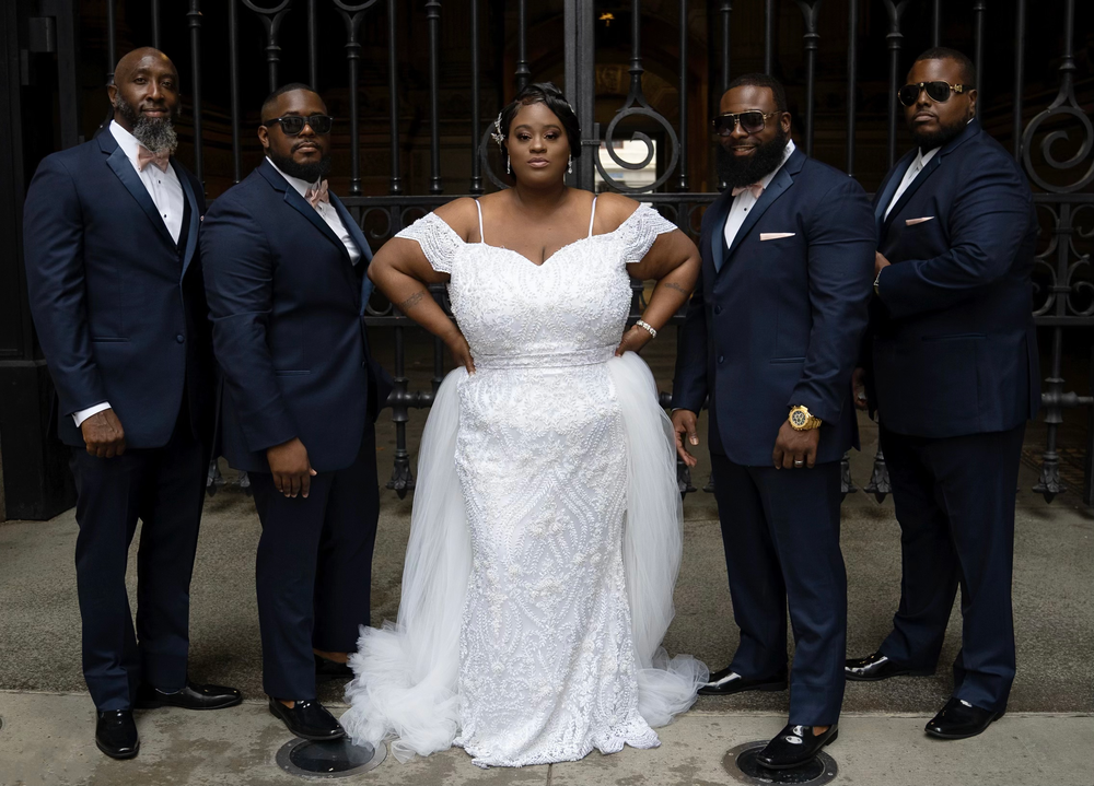 Wedding party posing in formal navy suits and white lace gown against ornate iron gates.