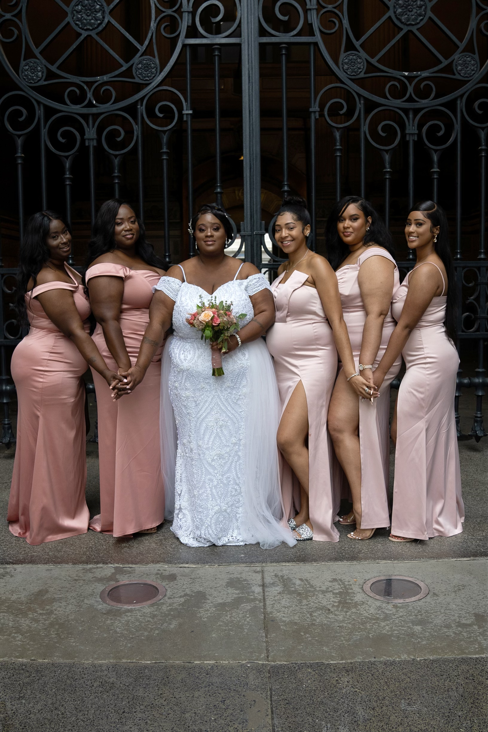 A bridal party poses together wearing pink dresses against an ornate iron gate.