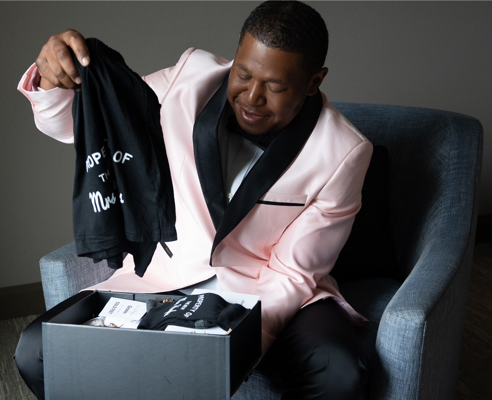 A person in a pink tuxedo jacket opening a gift box while seated in a gray chair.