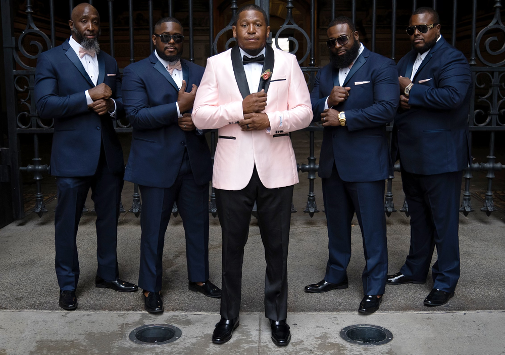 Groomsmen pose together in navy suits with one wearing a pink jacket in the center.