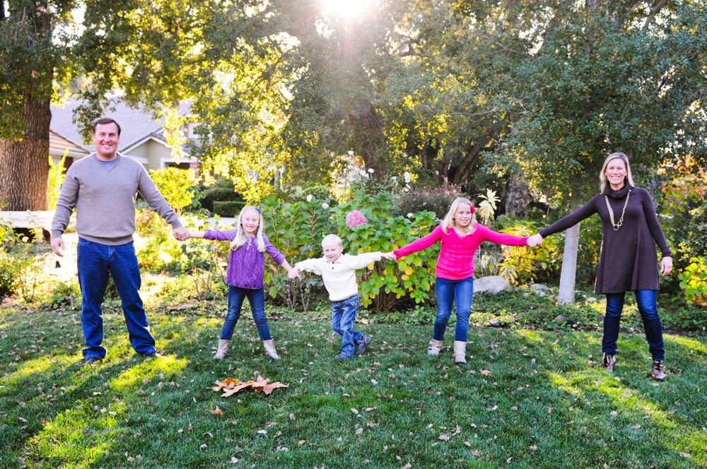 Family playing and jumping together on grass in a sunlit backyard garden setting.