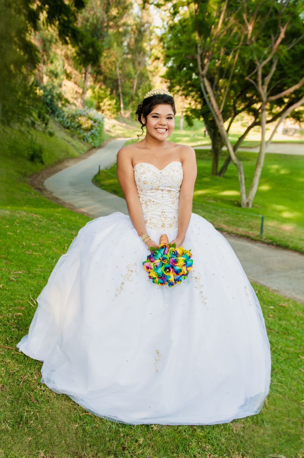Bride in stunning white ball gown holding colorful bouquet in park setting.