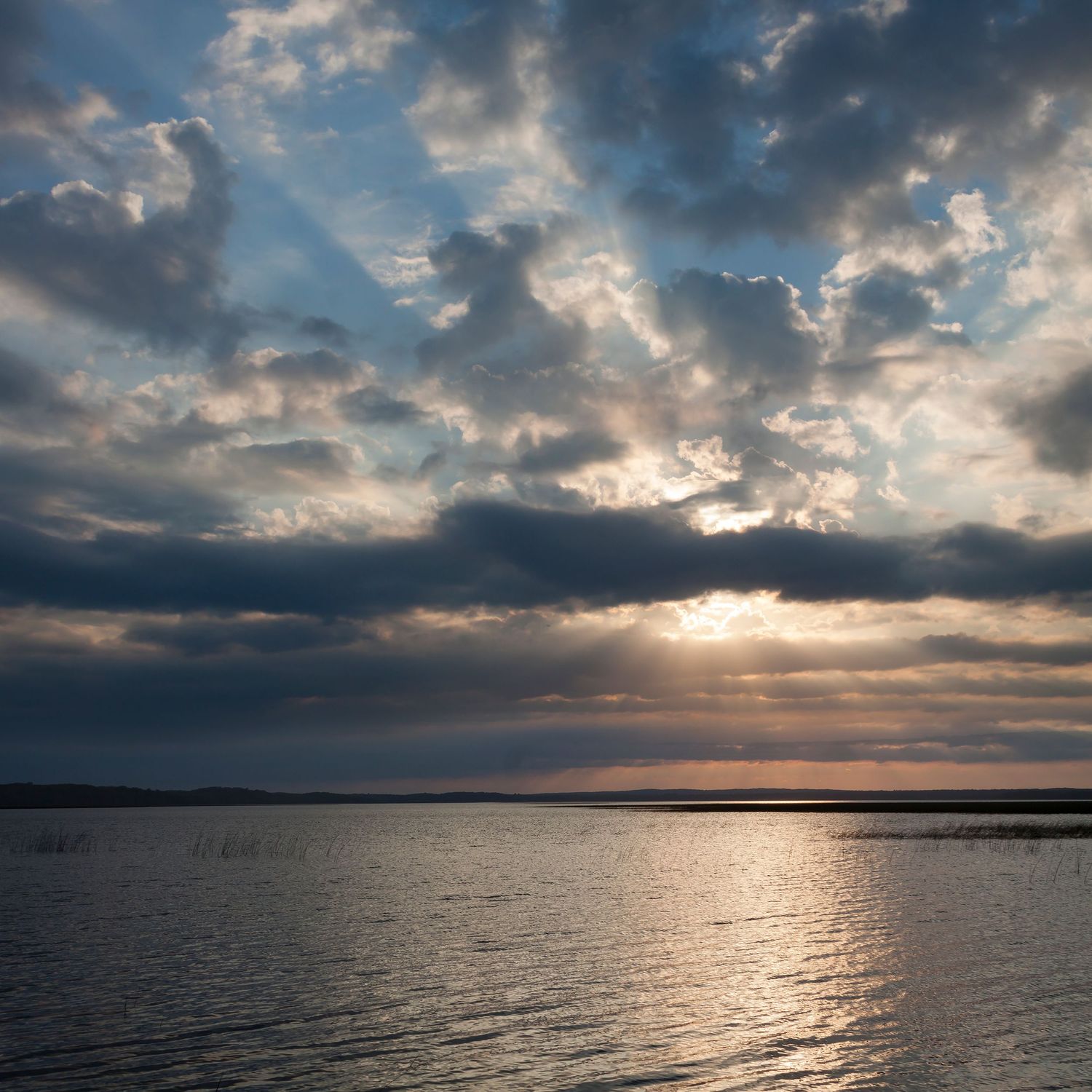 Dramatic sunbeams pierce through stormy clouds over calm water at sunset.