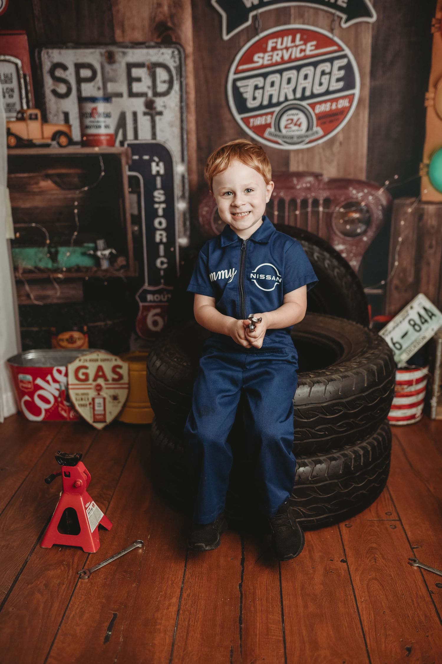 Young mechanic poses with tools and tires in vintage auto shop setting with retro signs and wooden floors.