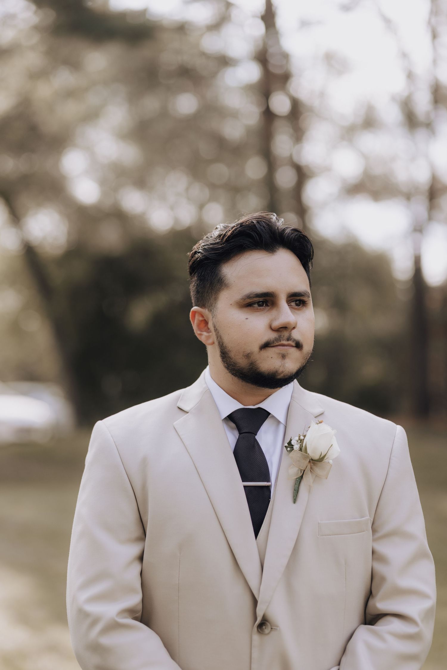 A person in a cream colored suit with black tie and boutonniere stands outdoors.