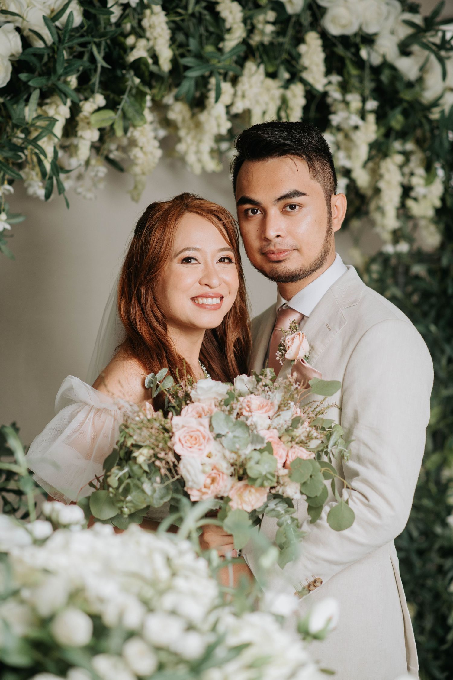 Elegant wedding portrait against lush white floral backdrop with soft natural lighting.