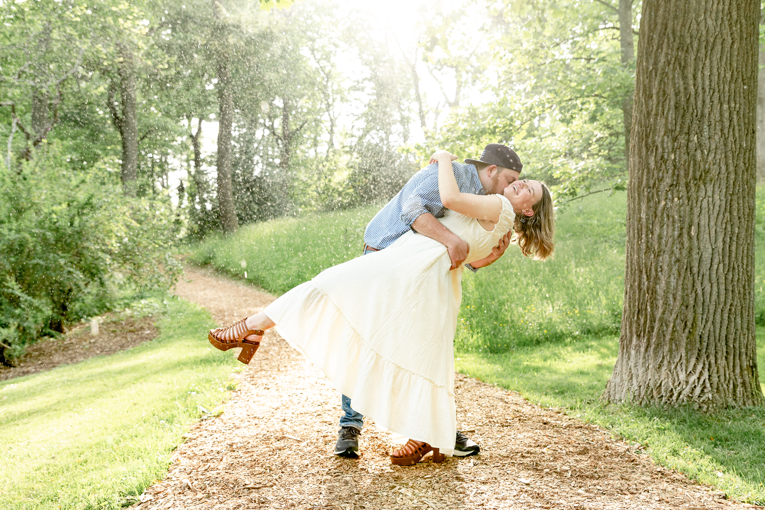 man in blue shirt dip woman in white dress under tree in rain with golden hour sun backlight