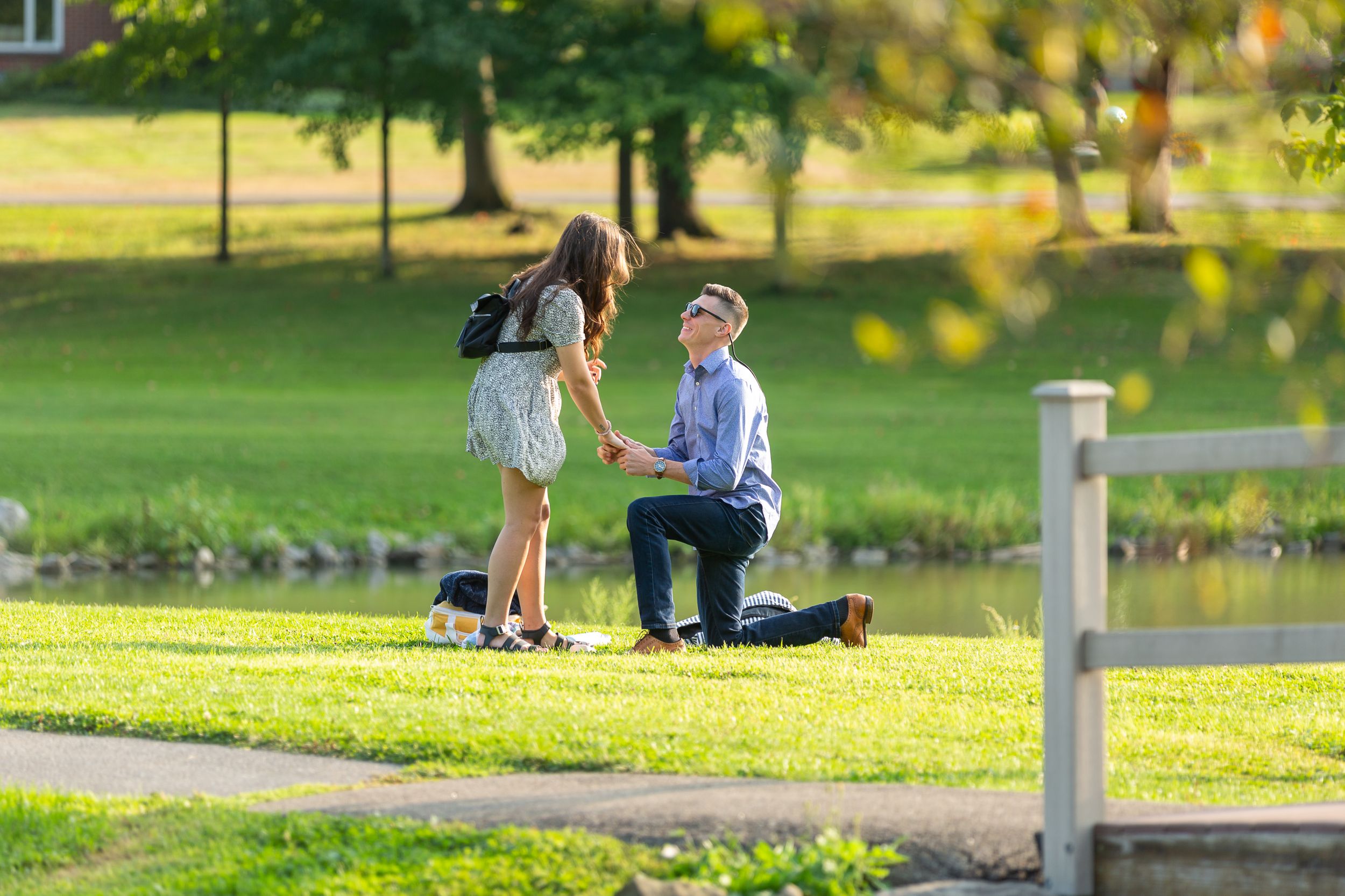 A couple shares a romantic proposal moment in a sunlit park by a lake.