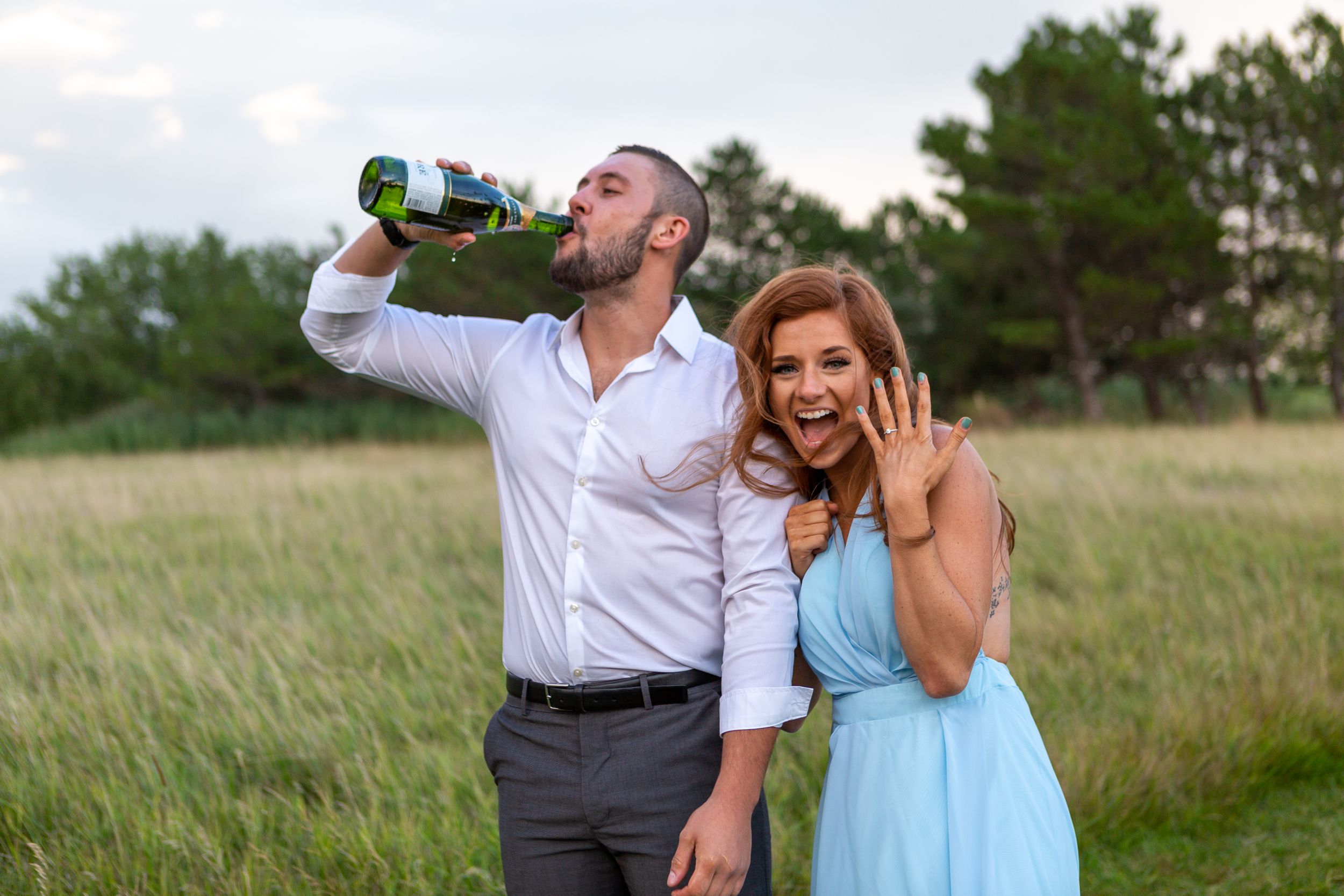 A couple celebrates with champagne in a grassy field while showing off an engagement ring.