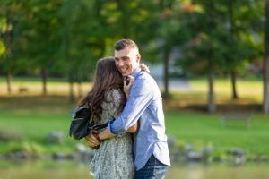A loving couple embraces in a park setting with trees and a lake in the background.