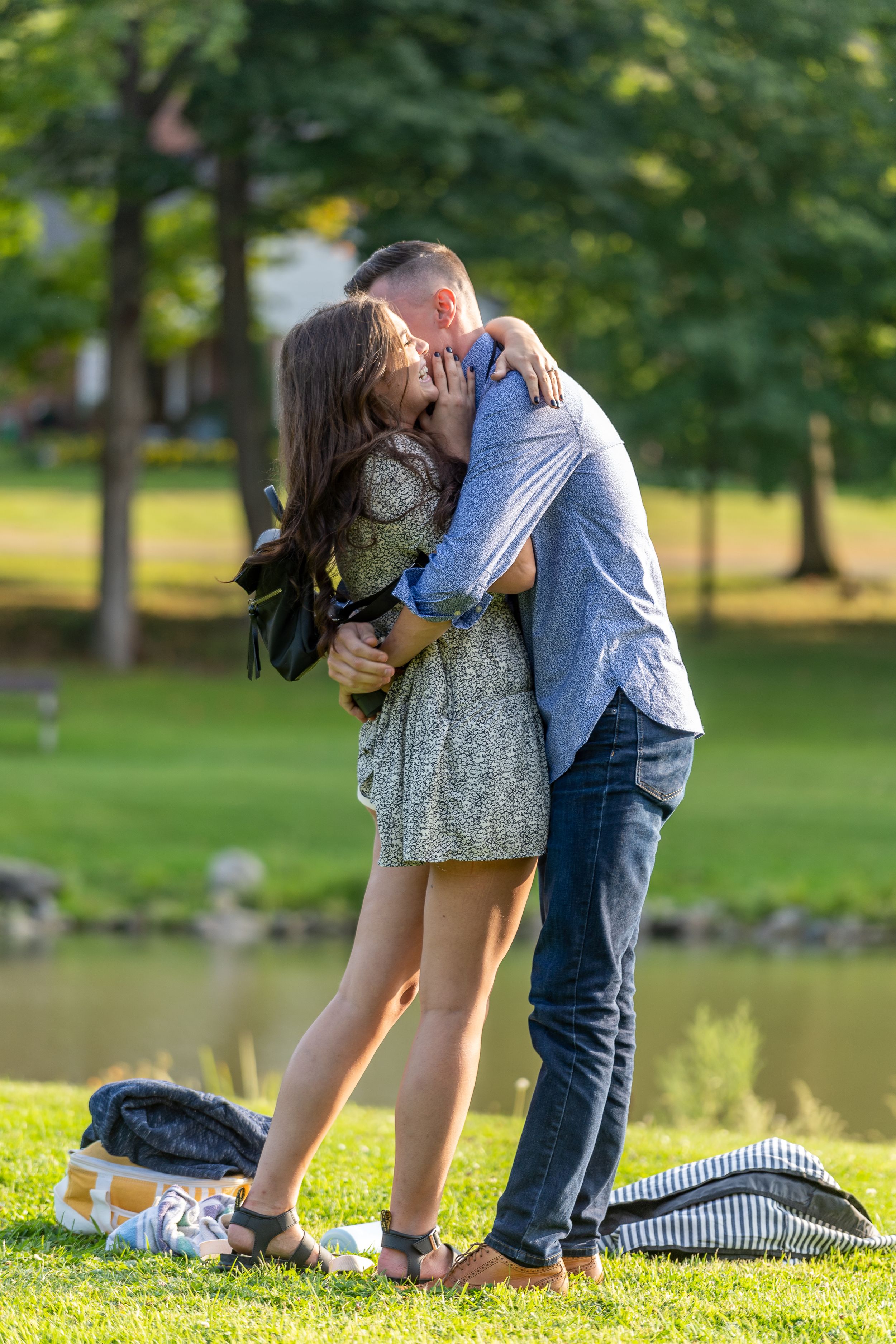 A couple shares a passionate kiss on grassy lakeside grounds during daytime.