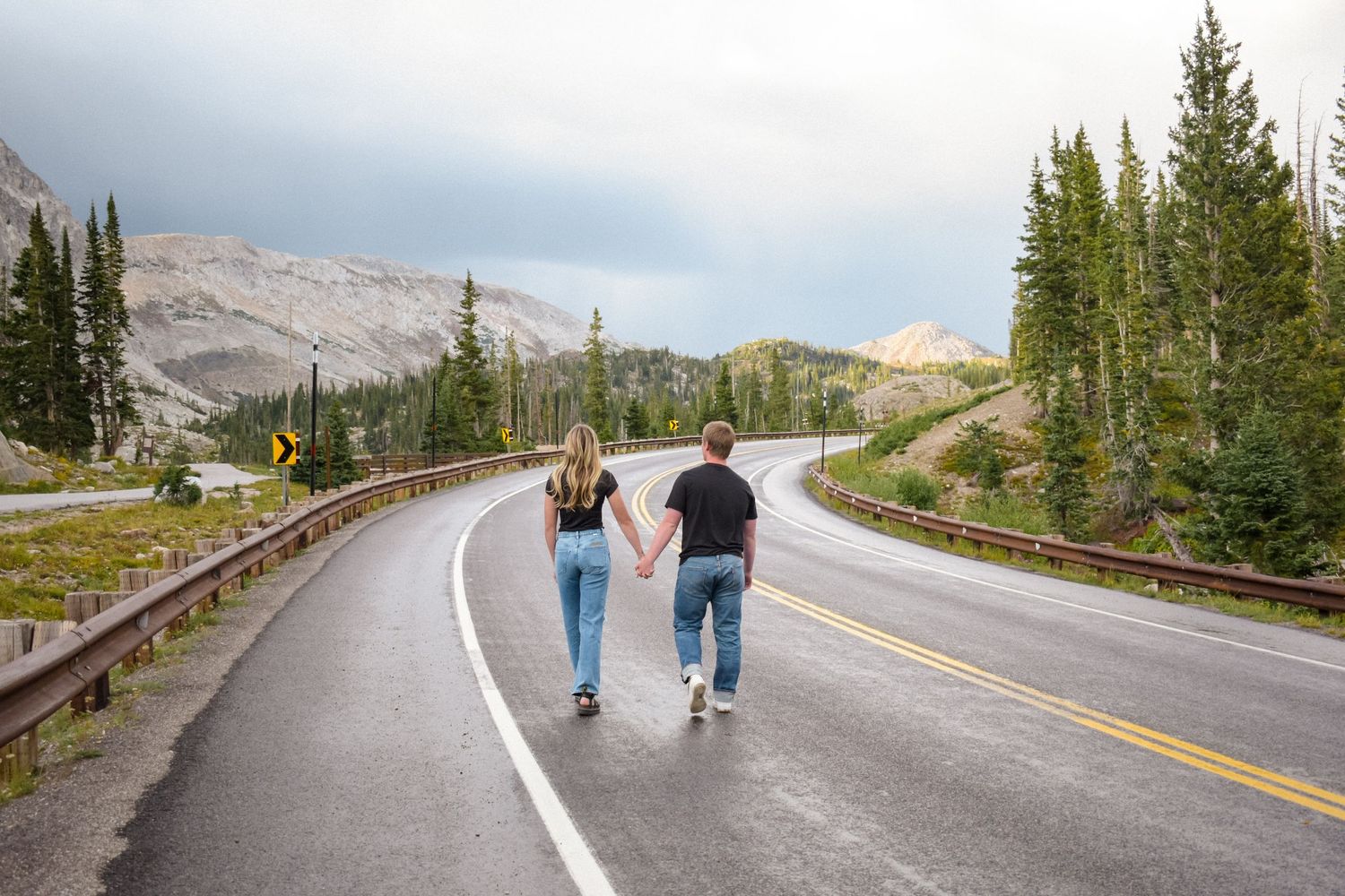 Two people walking hand in hand down a winding mountain road surrounded by evergreen trees.
