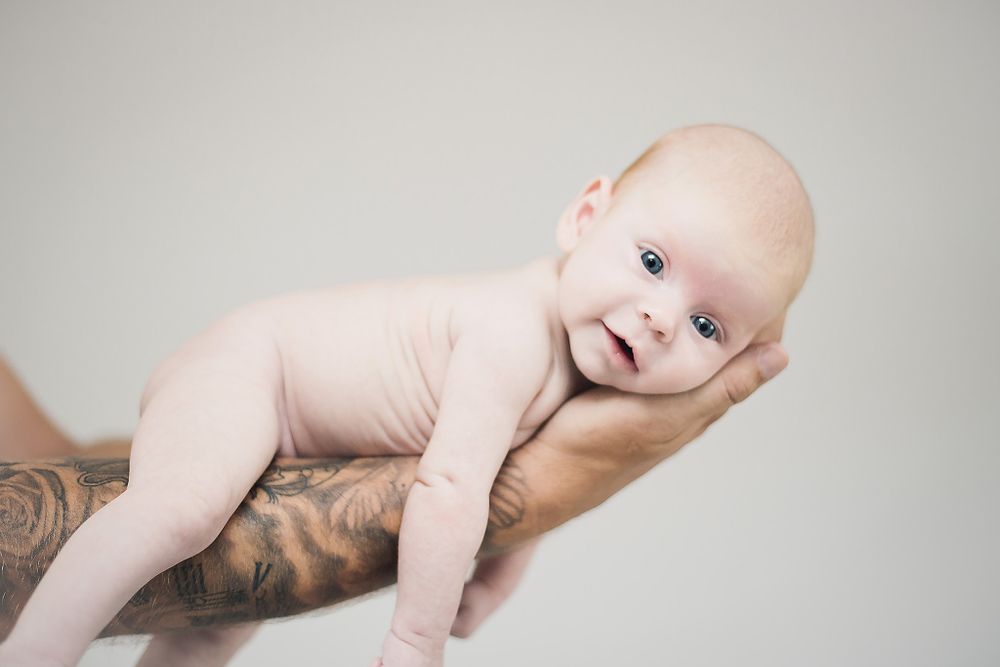 Infant resting on tattooed arm against light gray background.
