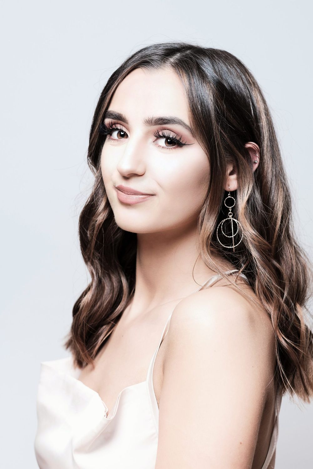 Woman in white top with wavy brown hair and statement earrings poses against light background.