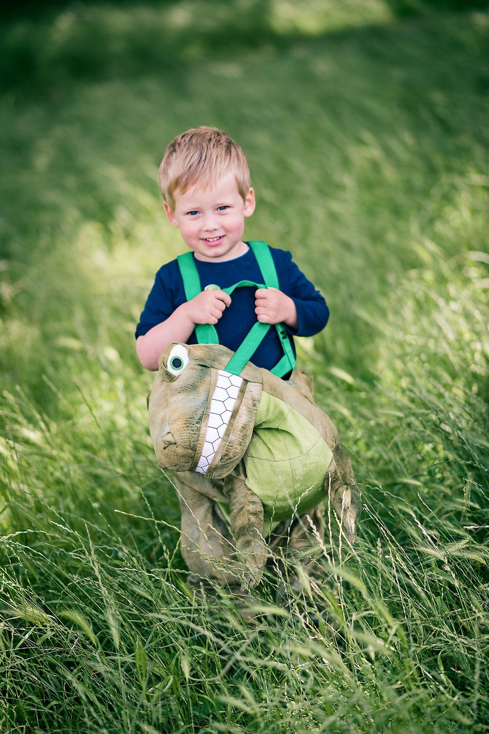Young child in green suspenders plays with a toy dinosaur in long grass.