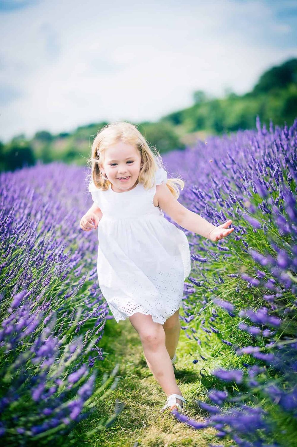 A child in a white dress runs joyfully through a blooming purple lavender field on a sunny day.