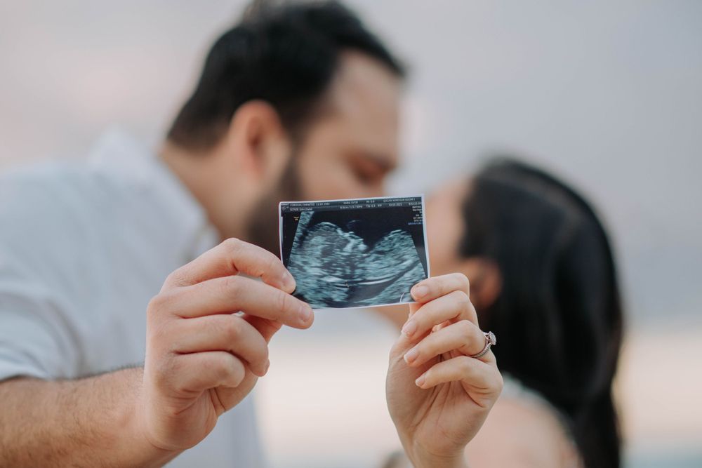A couple holds up their ultrasound photo against a sunset sky during a pregnancy announcement shoot.