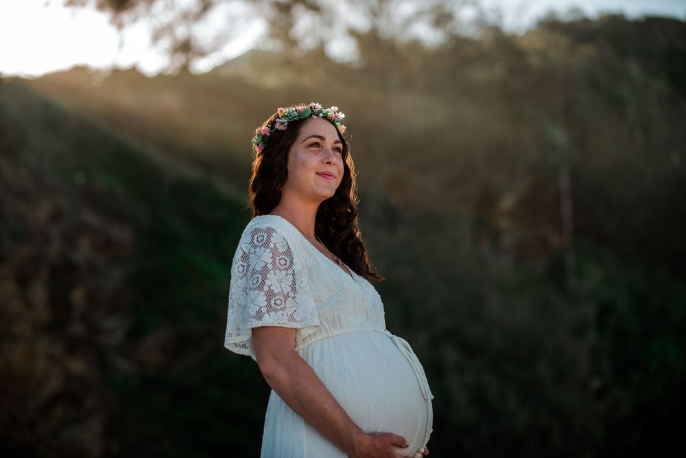 An expectant mother in a white lace dress stands in nature during sunset for a maternity photo.