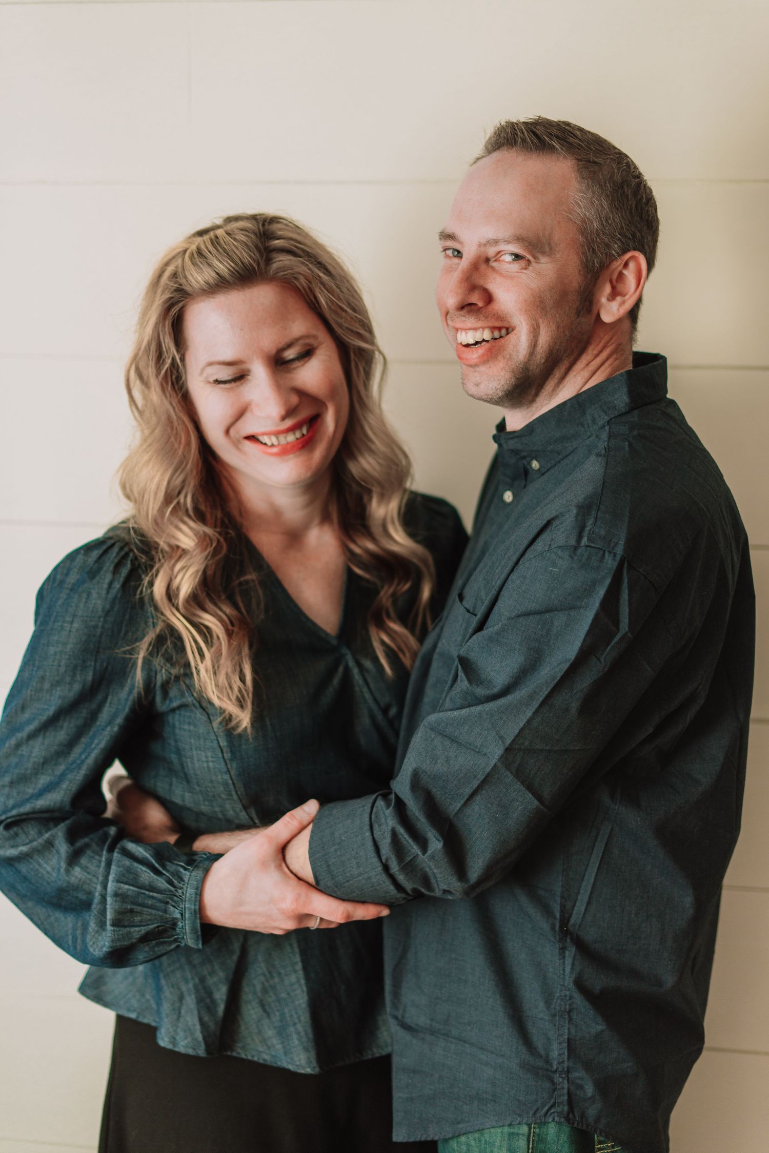 Couple sharing a joyful moment together while wearing coordinating dark outfits against a light wall.