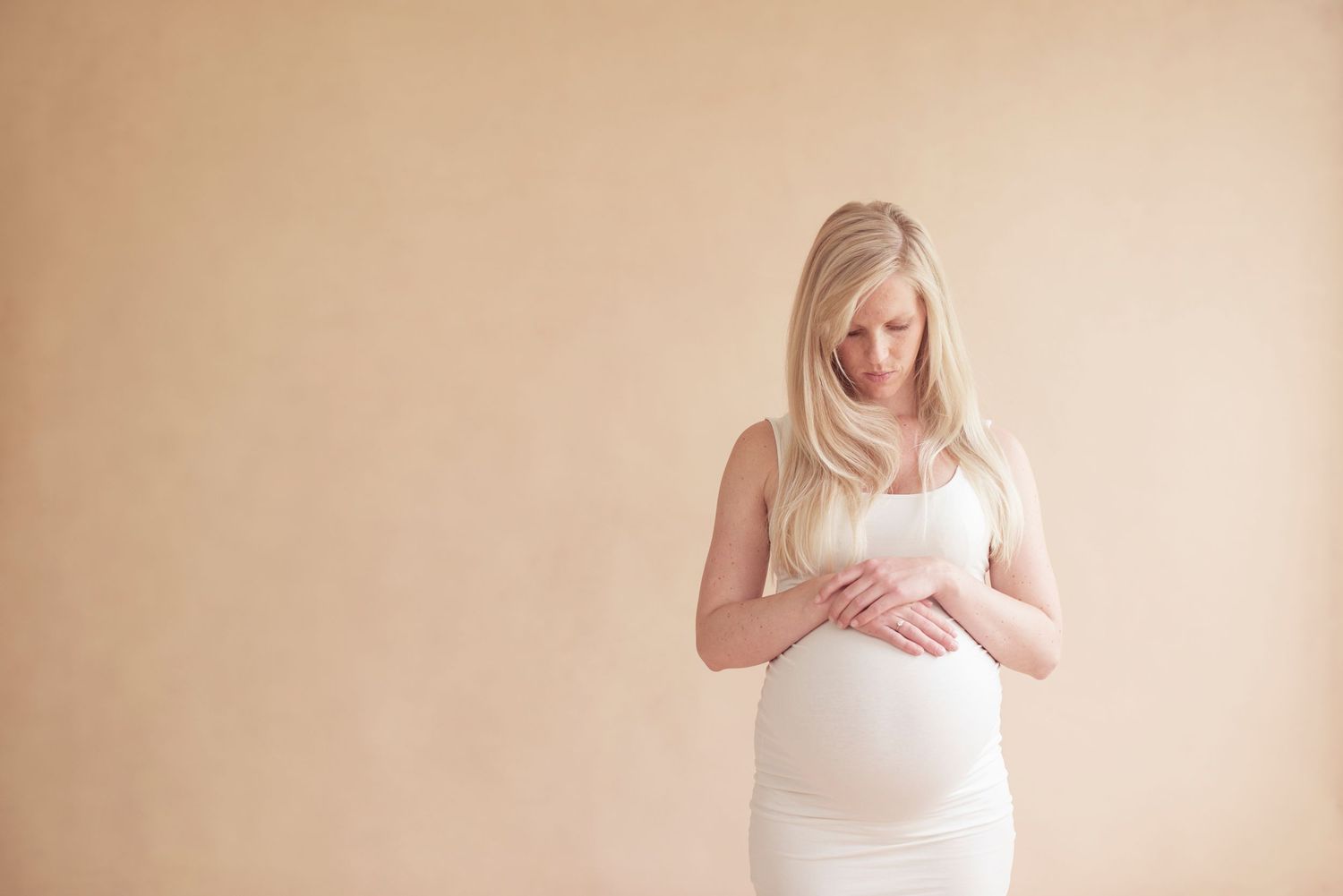 Pregnant woman in white dress gazes down at her baby bump against neutral background.