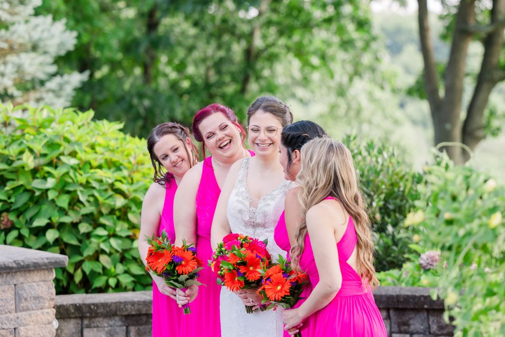 A bridal party in bright pink dresses holding vibrant red and orange bouquets in a garden setting.